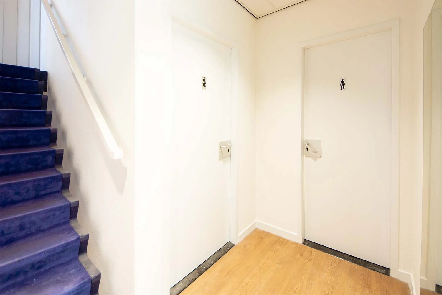 Gang met twee witte toiletdeuren voor dames en heren naast een trap met paarse bekleding in Huis-ter-Heideweg.